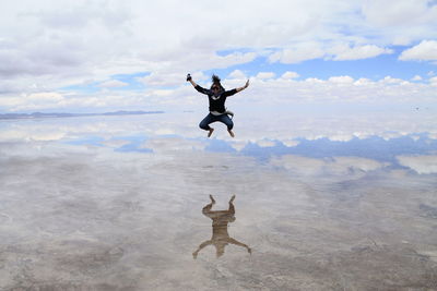 Woman in mid-air jumping over salt lake against cloudy sky at salar de uyuni