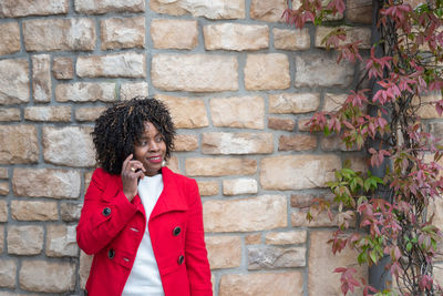 African american woman in red coat talking on smartphone near brick wall
