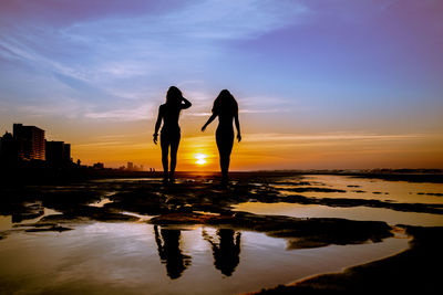 Silhouette women standing on shore against sky during sunset