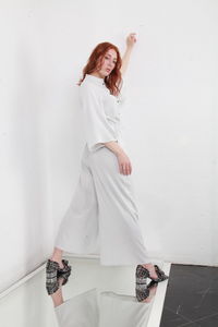 Back side view of fashion catalog style portrait of ginger woman in jumpsuit standing white wall