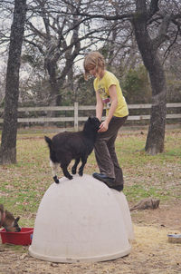 Girl and goat kid on white container against trees