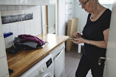 Senior woman standing in bathroom using mobile phone at home