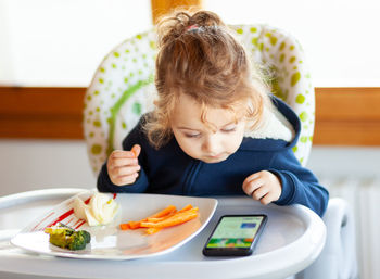 Cute girl eating and looking at mobile phone while sitting on seat at home