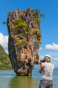 Rear view of woman photographing stack rock in sea against sky