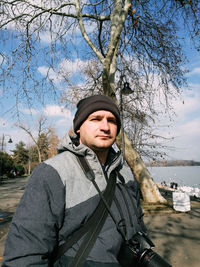 Portrait of young man with camera standing against bare tree and lake