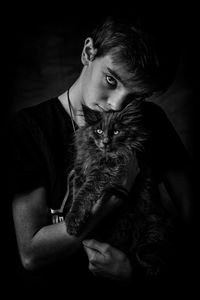Portrait of teenage boy carrying cat while standing against black background
