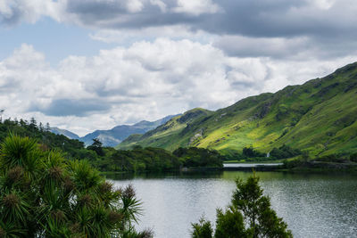 Scenic view of green mountains by lake against cloudy sky