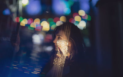 Woman taking selfie while holding illuminated string light in jar at night