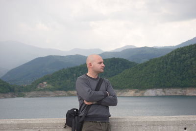 Thoughtful bald man with arms crossed standing against mountains