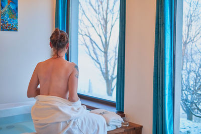 Rear view of shirtless woman looking through window at home