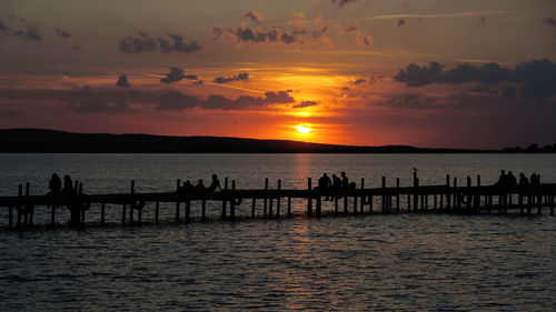 Silhouette people sitting on pier over sea against sky during sunset