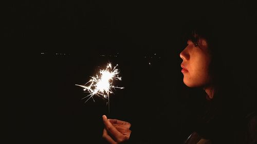 Side view of young woman holding fireworks against black background