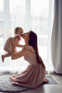 Young woman stands at a large window with a girl child in pink dresses at home barefoot