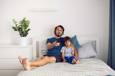 Bearded father and son are sitting on the bed in t-shirts and watching a football match on tv