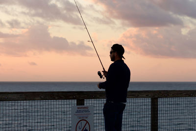 Rear view of man fishing by sea against sky during sunset