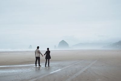 Rear view of couple holding hands while standing at beach during foggy weather