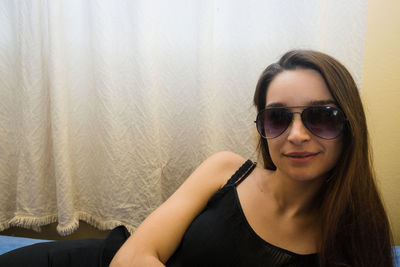 Portrait of young woman with long hair wearing sunglasses against curtain at home