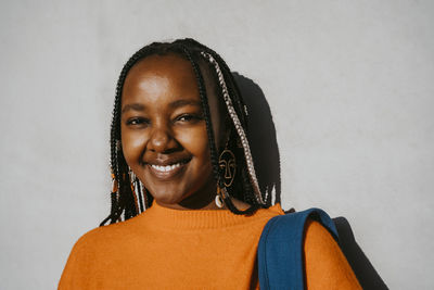 Portrait of smiling young woman with braided hairstyle against gray wall on sunny day