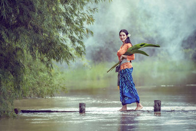 Full length of smiling woman carrying flowers while standing by lake