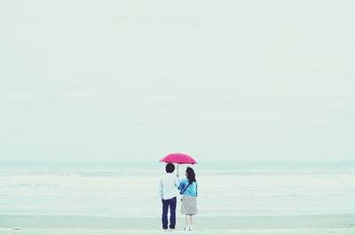 Rear view of friends with pink umbrella while standing at beach during rain