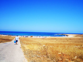 Rear view of man walking on road leading towards sea against clear blue sky during sunny day
