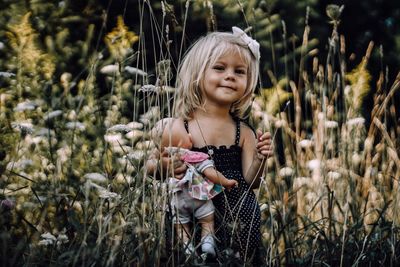 Portrait of innocent girl holding doll amidst plants on field