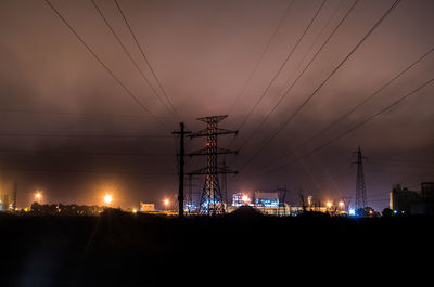 Low angle view of silhouette electricity pylon against cloudy sky in city at night