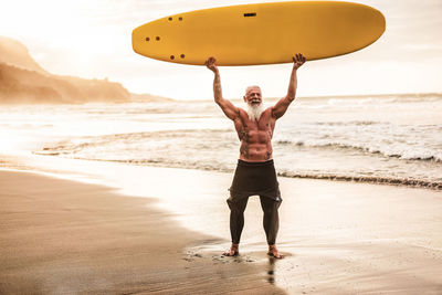 Full length of shirtless senior man holding surfboard at beach