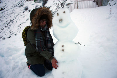 Young hiker kneeling on ground in the cold building a cute snowman in the white mountain in tuscany