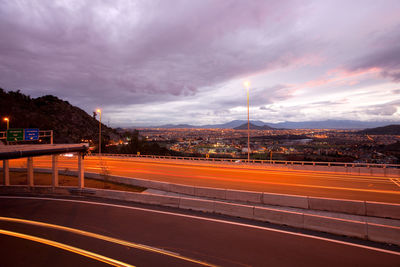 Light trails on road in illuminated city against cloudy sky at dusk