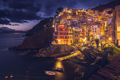 Illuminated city by sea against sky at sunset