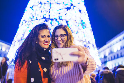 Smiling young women taking selfie while standing against illuminated lighting outdoors