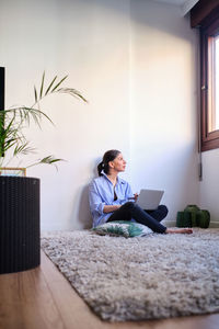 Concentrated barefoot female freelancer looking though window and typing on modern netbook while sitting on floor near white wall in light room