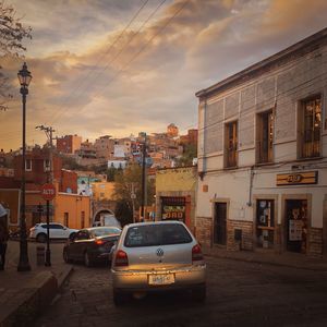 Cars on road by buildings against sky during sunset