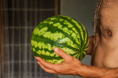 Midsection of shirtless man holding melon while standing at home