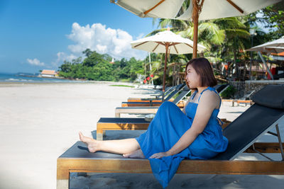 Young woman sitting on chair at beach against sky