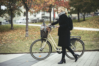Full length side view of businesswoman with bicycle walking on footpath during autumn