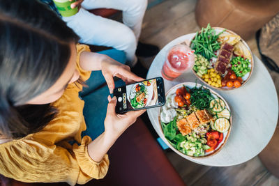 From above of anonymous female vlogger with black partner taking photo of healthy meal on cellphone in restaurant