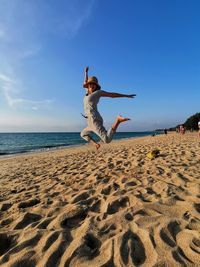 Full length of woman jumping on sand at beach against blue sky