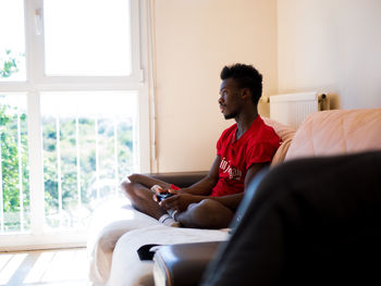 Young man using wireless playstation pad while sitting on bed at home