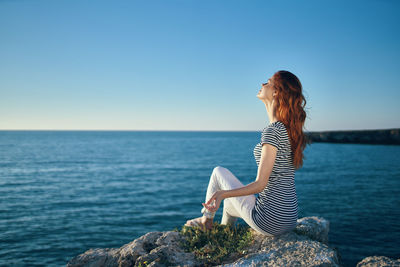 Woman sitting on rock by sea against clear sky