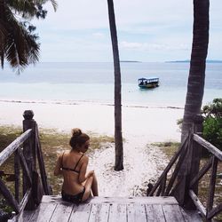 Rear view of woman sitting on stairs near beach against sky