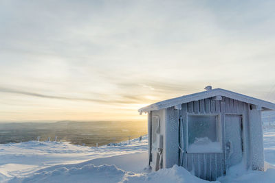 House on snow covered landscape against sky during sunset