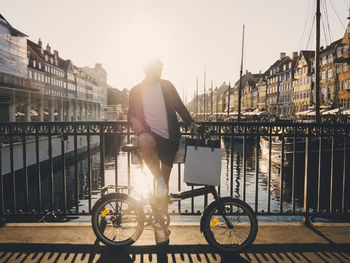 Full length back lit of man standing with bicycle and shopping bags on footbridge in city