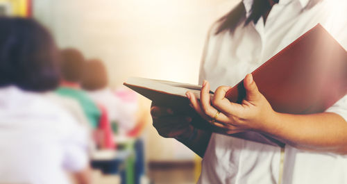 Midsection of teacher with book standing in classroom