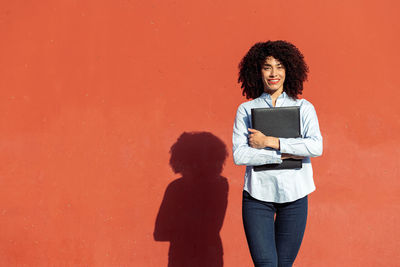 Full length of woman holding umbrella while standing against orange wall