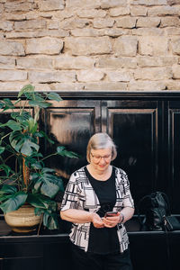 Senior female tourist using smart phone while standing against stone wall at restaurant