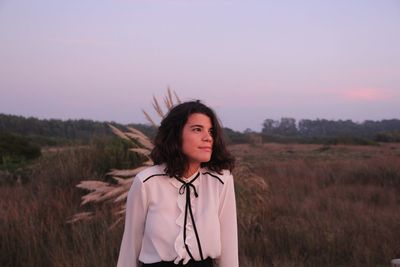 Beautiful young woman looking away while standing on field against sky during sunset