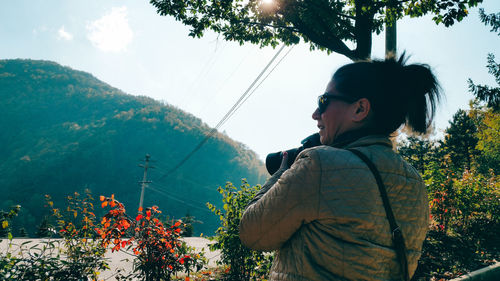 Side view of woman with camera standing by tree against sky