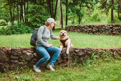 A girl with a backpack is sitting on the lawn stroking a labrador retriever and smiling 